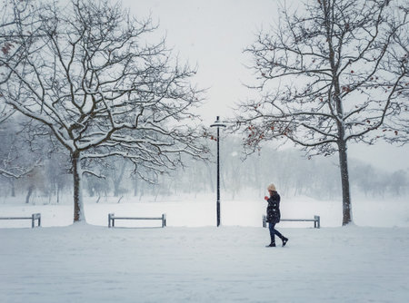 Lone young woman walking in the park drinking a hot coffee in a cold winter day. Seasonal blizzard weather on the street. Snowfall morning in the square. Gils wandering under snowy treesの写真素材