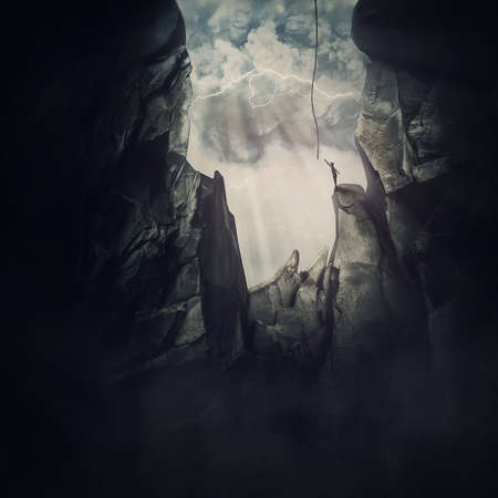 Conceptual scene with a man escaping from the steep chasm, stretching to reach the helping rope and get out of the darkness. Big cave inside the rocky mountain and person trying to break freeの写真素材