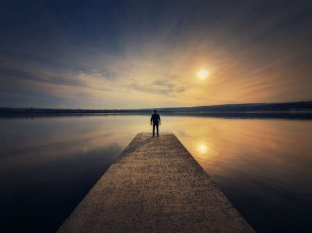Man standing alone on the pier facing the sunset reflected in the calm lake water. Scenic view with a lonely person on the dock looking at the horizonの写真素材