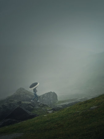 Man with umbrella climbing a mountain hill in a gloomy weather with dense mist. Moody hiking scene with a wanderer walking on the cliffs of the foggy valleyの写真素材