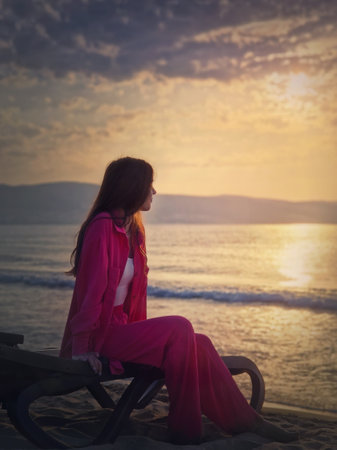 Carefree young woman aesthetic portrait sitting relaxed on the sunbed at the beach looking at the sunrise above the sea. Beautiful seaside dawn scene. Summer holiday, travel and relaxation conceptの写真素材
