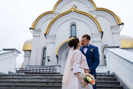 the bride and groom on the background of a church, religious lovers, a wedding walk in winter, a man and a woman gently kiss and smile hugsの写真素材