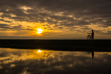 Silhouette of a boy driving his bicycle along the seashore at sunset.A narrow strip of land, in the water reflects a yellow sunset, clouds and a boy with a bicycle.の写真素材