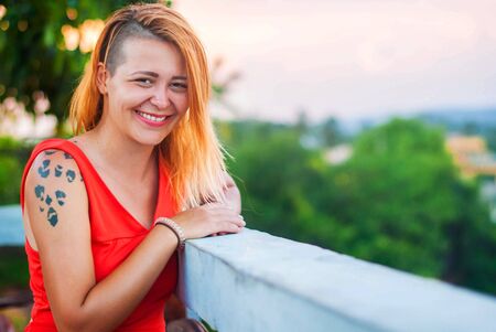 Beautiful red-haired girl in a red dress and tattooed laughs on the veranda of a summer restaurantの写真素材