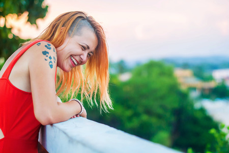 Beautiful red-haired girl in a red dress and tattooed laughs on the veranda of a summer restaurantの写真素材