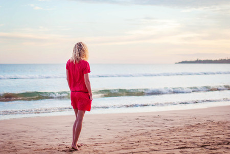 Beautiful romantic girl blonde in a red dress on the ocean shore during sunset. Silhouette of a girl on a sunset background near the ocean. Beautiful girl on the beach in a red dress.の写真素材