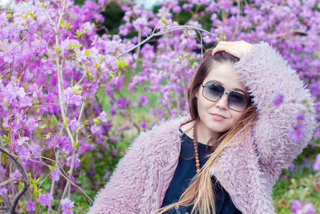 Beautiful girl in an artificial pink coat. Girl with long hair wearing sunglasses. The woman smiles in the sunglasses and pink coat in the magnolia garden.の写真素材