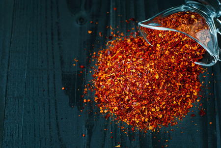Ground chilli pepper scattered on a dark wooden table. Spice chili on a dark background. Dried chile Pieces. Spice in a glass vase.の写真素材