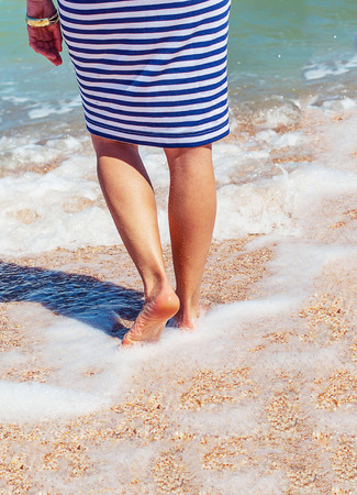 Tanned legs of a girl with a red pedicure on the beach. Female legs in striped dress in the sea. Legs of a woman with a red pedicure in the sea.の写真素材