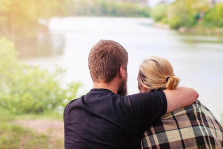 A man and a woman in a rug sit embracing on the river bank. A loving couple hugs in a rug. A guy with a beard and a blonde girl are sitting hugging.の写真素材