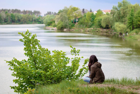 A young girl with long dark hair in a cloak with a hood sits on the river bank with her back and looks afar at nature.の写真素材