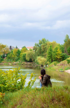 A young girl with long dark hair in a cloak with a hood sits on the river bank with her back and looks afar at nature.の写真素材