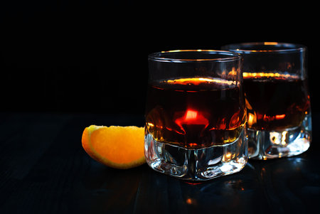 Two glasses of whiskey or cognac with an orange slice on a black background and a wooden tableの写真素材