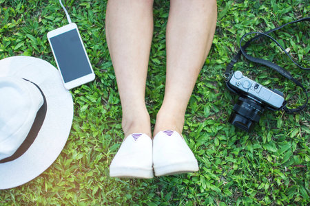 Top view  legs of Asain tourist woman wear white shoes sitting for relaxing with white hat, camera and smartphone on a green grass at the park, Hobby free time and leisure timeの写真素材