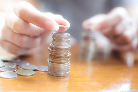 Hand of woman putting  a  silver coin on top of two pile for stacking on the wooden table, selective...