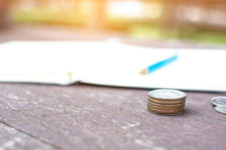 Close up rising coins pile of money coins stack and pencil lied on a notebook on the wooden table with bokeh of green garden backgroundの写真素材