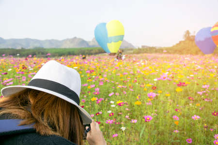 Tourist woman take a shot of photo by digital camera to beautiful blooming cosmos flower garden with with hot air balloon in a field, photography lifestyleの写真素材