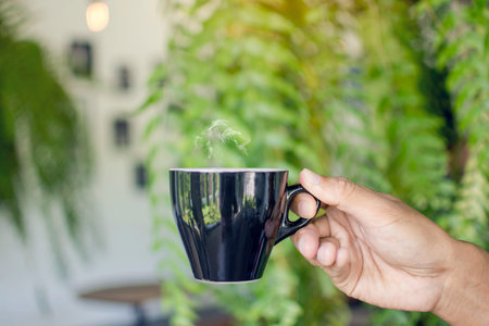 Asian man hand holding black cup of hot coffee with steaming on blurred leaves