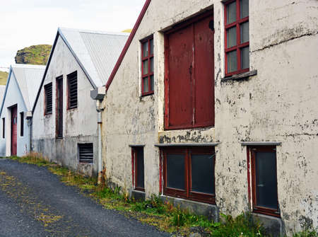 A row of disused industrial buildings stand at the side of a narrow track on the island of Heimaey, Iceland. Once used as industrial and storage units most of the buildings are now empty and unusedの写真素材