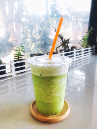 Green tea milk frappe in clear plastic glass have orange straw in wooden tray put on dining table with cactus, succulent and mirror.の写真素材