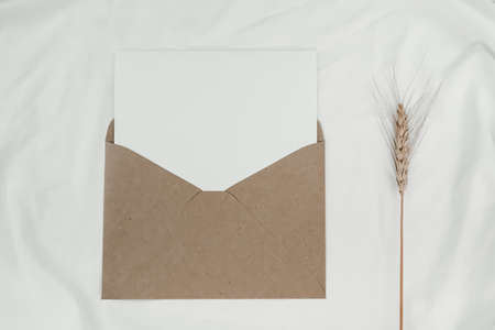 Blank white paper is placed on the open brown paper envelope with Barley dry flower on white cloth. Mock-up of horizontal blank greeting card. Top view of Craft paper envelope on white background. Flat lay of stationeryの写真素材