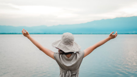 Young asian woman wearing a hat and raising her arms outstretched with lakes and mountains....