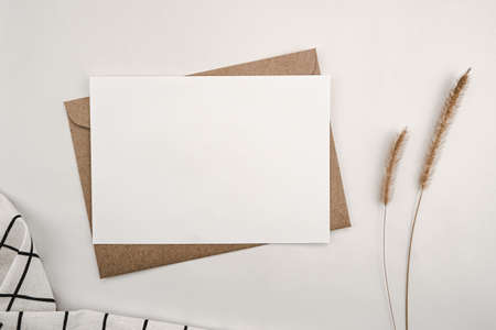 Blank white paper on brown paper envelope with Bristly foxtail dry flower and White cloth with Black grid pattern. Mock-up of horizontal blank greeting card. Top view of Craft envelope on white background.の写真素材