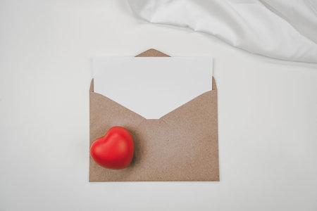 Blank white paper is placed on open brown paper envelope with red heart, White cloth. Top view of...