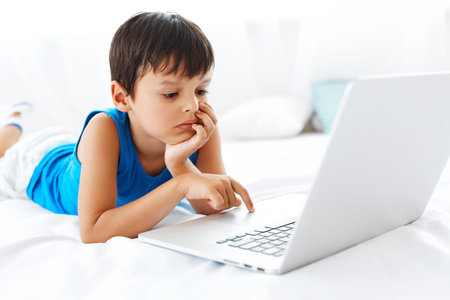 Young boy using notebook in his bedroom. Child is typing words while lying on the bed.