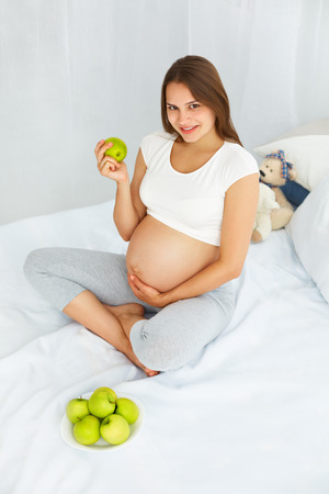 Pregnant Young Woman holding Apple while sitting on the Bed. Healthy Food and Diet Concept. Healthy Lifestyle.の写真素材