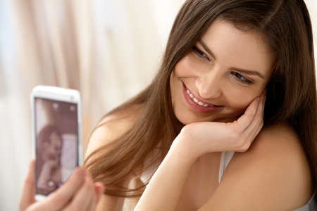 Portrait of a Young Woman Holding a Smartphone Digital Camera and Taking a Selfieの写真素材