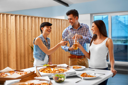 Happy Friends Having Fun At Dinner Party At Home. People Eating Pizza, Drinking Beer And Celebrating Holiday Indoors. Friendship, Leisure, Celebration Concept. Fast Food.の写真素材