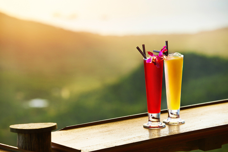Drinks. Close Up Of Exotic Cocktails In Tall Glasses With Orchids And Drinking Straws On Wooden Table In Luxury Tropical Bar.  Blur Thai Hills, Sky On Background. Thailand Vacations. Celebration.の写真素材
