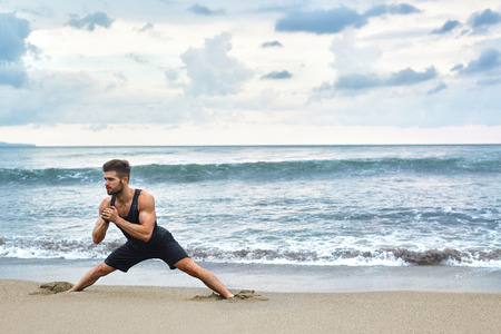 Fitness Exercise. Sporty Handsome Male Stretching Legs Before Run. Fit Athletic Man With Muscular Body Exercising On Sand, Training During Outdoor Workout At Beach. Sports, Healthy Lifestyle Conceptの写真素材