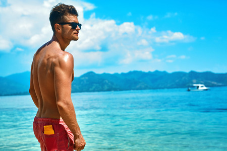 Summer Travel Vacation. Handsome Man With Sexy Body In Fashionable Sunglasses Sunbathing, Tanning At Sea Beach. Fitness Male Model With Sunscreen Lotion, Sun Block Skin Protection Cream In Pocketの写真素材