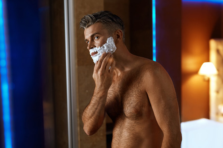Man Using Shaving Cream On His Face In Fashionable Luxury Bathroom. Closeup Portrait Of Male Model With Naked Body Applying Foam Before Shaving Beard. Men Facial Hair Grooming, Skin Care Conceptの写真素材