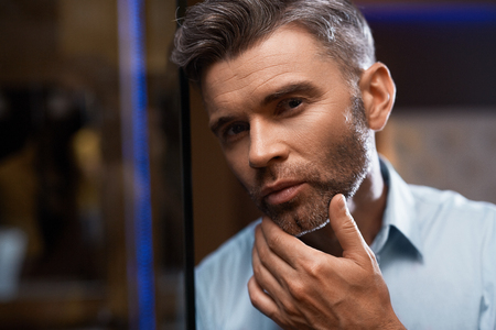 Men Grooming. Handsome Man With Stubble Touching Face Or Beard Looking Into Mirror. Closeup Portrait Of Successful Confident Male Model In Elegant Fashion Clothes In Luxury Interior. Skin Care Conceptの写真素材