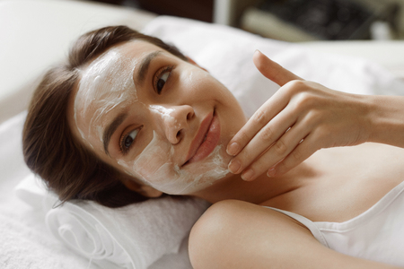 Face Skin Care. Closeup Of Beautiful Healthy Smiling Woman With White Cosmetic Mask On Her Face Relaxing And Enjoying Facial Beauty Treatment At Day Spa Salon Indoors. High Resolution Imageの写真素材