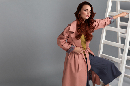 Beautiful Fashion Model Wearing Fashionable Clothes On Grey Background. Portrait Of Sexy Woman In Stylish Glamour Style Clothing, Light Pink Coat, Dark Purple Trousers Posing On White Ladder In Studioの写真素材