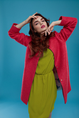 Fashion Model Girl In High Fashionable Clothing Posing On Blue Background. Beautiful Sexy Woman With Perfect Makeup And Elegant Hairstyle Wearing Trendy Clothes, Green Dress, Stylish Dark Pink Coatの写真素材