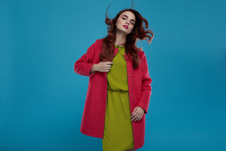 Fashion Model Girl In High Fashionable Clothing Posing On Blue Background. Beautiful Sexy Woman With Perfect Makeup And Elegant Hairstyle Wearing Trendy Clothes, Green Dress, Stylish Dark Pink Coatの写真素材