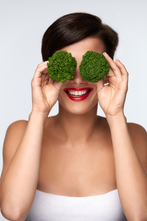 Food For Health. Beautiful Smiling Woman With Red Lips Holding Organic Green Broccoli Covering Her Eyes. Portrait Of Happy Girl Playing With Vegetable. Healthy Diet Nutrition Concept. High Resolutionの写真素材