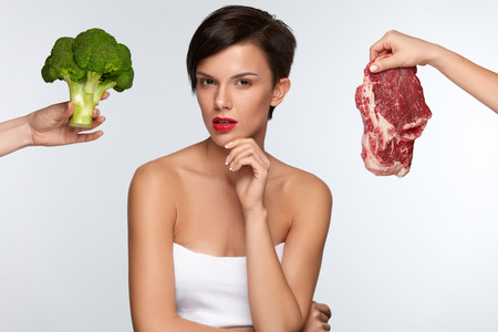Healthy Eating. Beautiful Woman With Red Lips Choosing Between Raw Meat And Broccoli, Thinking Whether To Eat Vegetables Or Meat. Female Hands Holding Food Products. Nutrition Concept. High Resolutionの写真素材