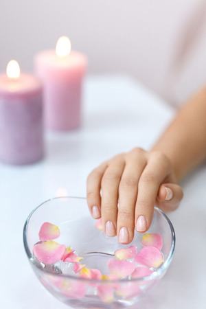 Spa Procedure. Woman In Beauty Salon Holding Fingers In Aroma Bath For Hands. Closeup Of Female Nails Soaking In Bowl Of Water With Floating Pink Flower Petals. Skin Care. High Resolutionの写真素材