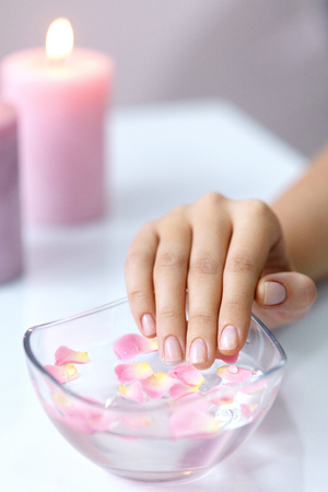 Spa Procedure. Woman In Beauty Salon Holding Fingers In Aroma Bath For Hands. Closeup Of Female Nails Soaking In Bowl Of Water With Floating Pink Flower Petals. Skin Care. High Resolutionの写真素材