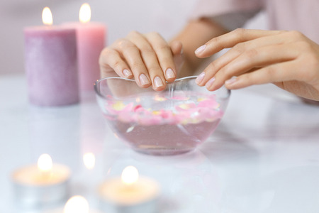 Spa Procedure. Woman In Beauty Salon Holding Fingers In Aroma Bath For Hands. Closeup Of Female Nails Soaking In Bowl Of Water With Pink Flower Petals. Aromatherapy. High Resolutionの写真素材