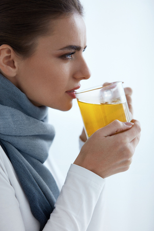 Beautiful Sick Woman Drinking Medicine From Cold And Flu. Closeup Of Young Female In Scarf Drinking Hot Tea. Portrait Of Girl Feeling Cold, Holding Big Cup Of Yellow Warm Drink. High Resolutionの写真素材