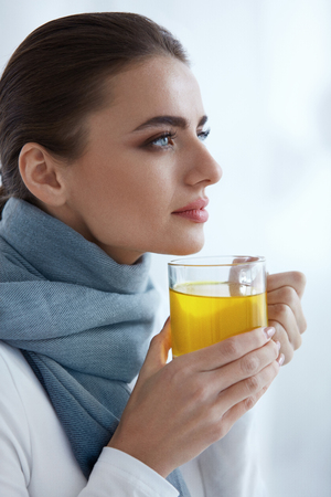Beautiful Woman In Warm Scarf Drinking Hot Yellow Drink. Closeup Of Smiling Young Female Holding Cup Of Warming Tea, Liquid Medicine From Cold And Flu In Hands Indoors. Health Concept. High Resolutionの写真素材