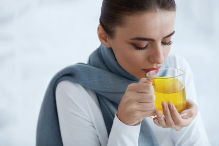 Cold. Closeup Of Beautiful Young Woman In Warm Scarf With Hot Cup Of Tea In Hands. Sickness And Illness. Portrait Of Sick Unhealthy Female Drinking Warming Yellow Drink Indoors. High Resolutionの写真素材