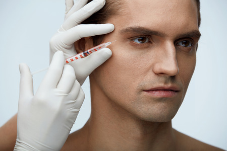 Facial Beauty. Portrait Of Handsome Man Getting Hyaluronic Collagen Injection. Closeup Of Beautician Hands Holding Syringe With Transparent Liquid Near Male Face. Skin Lifting. High Resolutionの写真素材